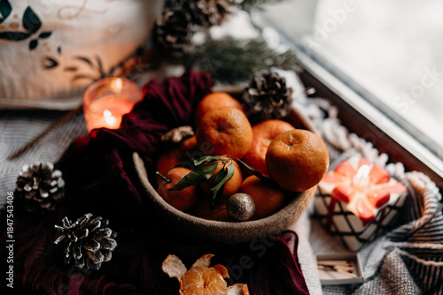 Winter still life by the window with tangerines, pine cones and candle. Cozy festive scene with natural daylight and calm home comfort mood.
