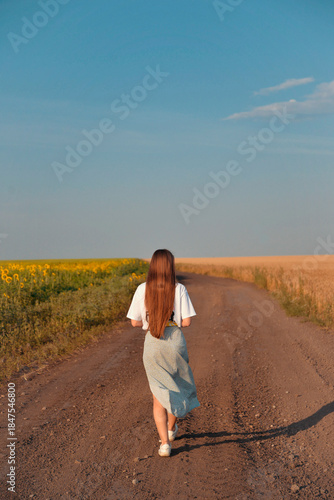 A young woman with long brown hair walks along a dirt road, bordered by a field of sunflowers and a grain field.