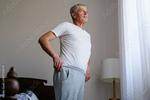 An elderly man stands thoughtfully by a window in his bedroom, dressed in a white shirt and grey pants, absorbing the morning light