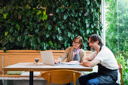 Tableau sur toile Two colleagues, cafe owner and financial advisor, in a cafe with a laptop discuss work beside a lush indoor green wall, suggesting consulting or small business planning in a modern, casual workspace