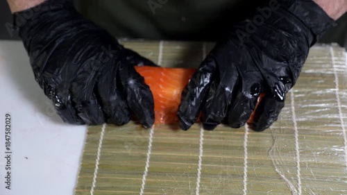 A chef wearing gloves prepares sushi rolls with salmon on a bamboo mat in a professional kitchen. The sushi cooking process. Asian cuisine, cooking process.