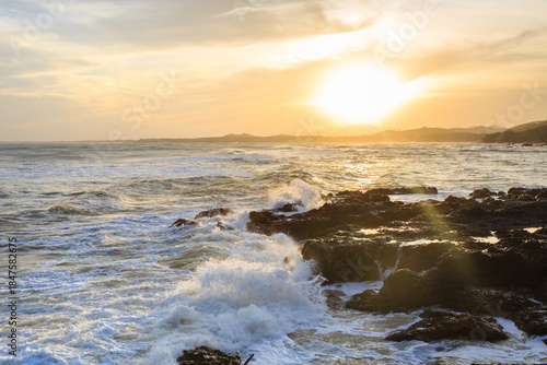 Backlit, the surging sea crashes against the black rocks, splashing up white spray.