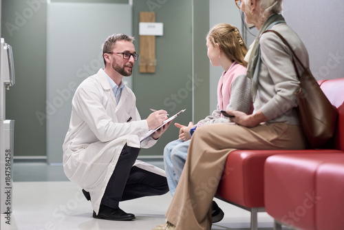 Caucasian male doctor kneeling and speaking with Caucasian girl child while senior Caucasian woman sitting nearby in waiting area, doctor holding clipboard and pen, medical consultation scene