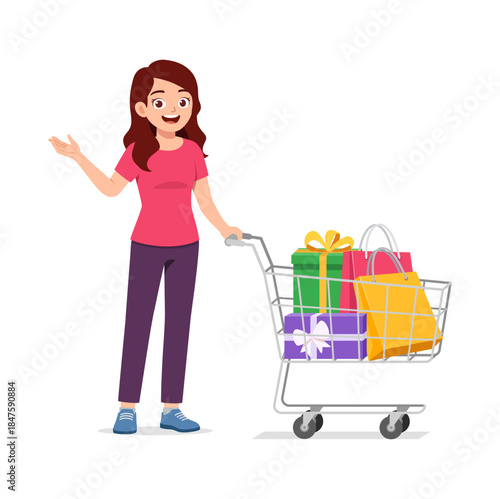 Happy young woman pushing shopping cart with gifts and bags smiling widely and gesturing with open hand