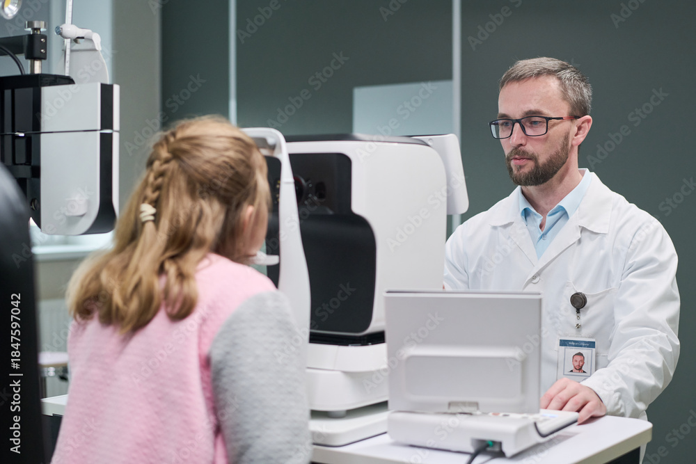 Obraz premium Caucasian middle aged man optometrist conducting eye examination for Caucasian child girl using modern ophthalmic equipment in medical clinic, both focused on diagnostic process