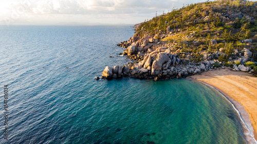 Aerial view of the vivid turquoise waters meeting the golden sands of a secluded beach beneath rocky outcrops and tree-covered headland, Magnetic Island, Queensland, Australia.