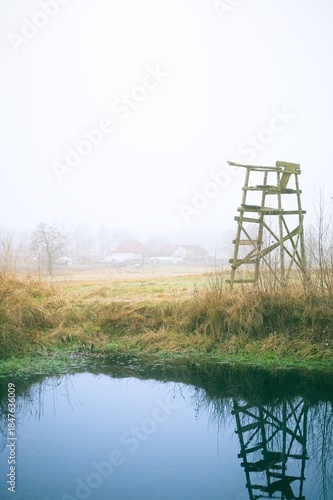 Autumn landscape of the Polish countryside in dense fog. A wooden hunting stand reflects in a small pond. Rural buildings in the distance. Calm, melancholy, and raw nature in pastel tones.