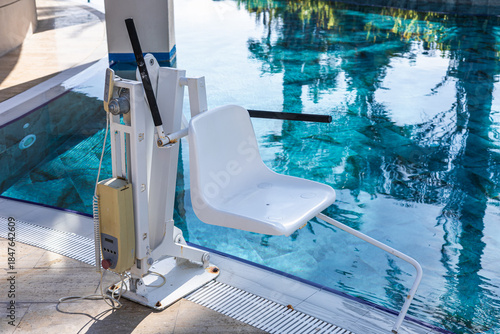 A poolside scene featuring a white pool lift beside a clear blue swimming pool. Lush greenery and a wooden bridge are visible in the background.