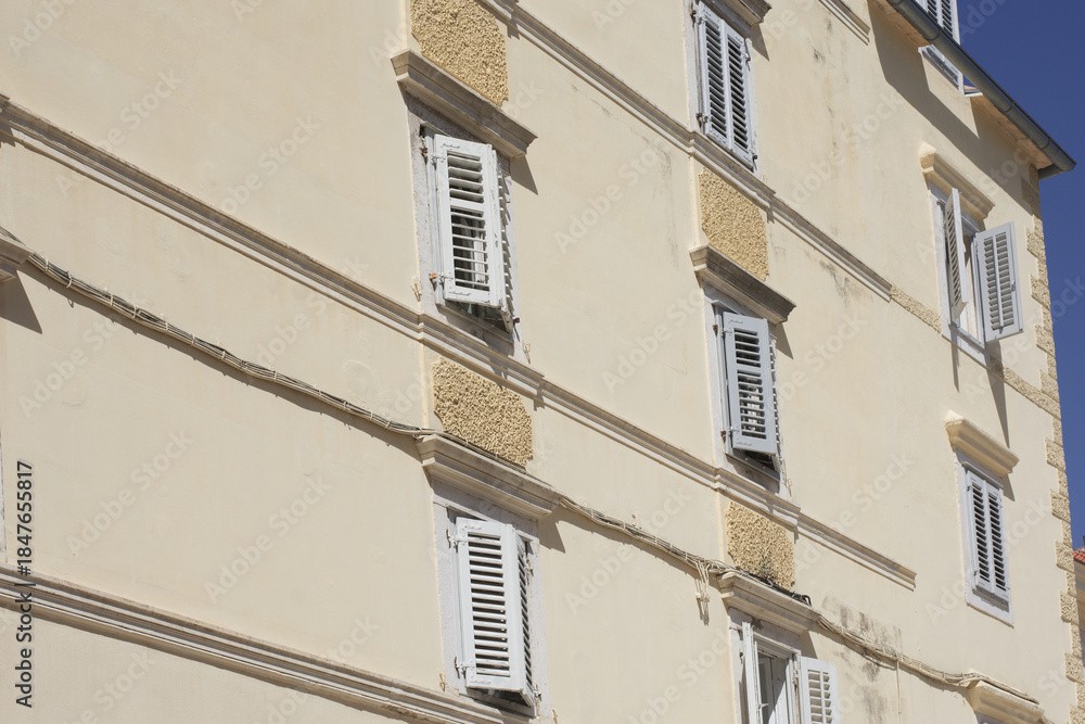 Obraz premium Two simple, square windows with white frames set into a weathered, rustic stone and plaster wall of an old European building under a terracotta roof.