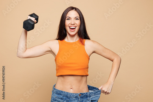 Young woman wear orange casual clothes hold dumbbell show loose denim pants after weight loss isolated on plain pastel beige background. Proper nutrition healthy fast food unhealthy choice concept