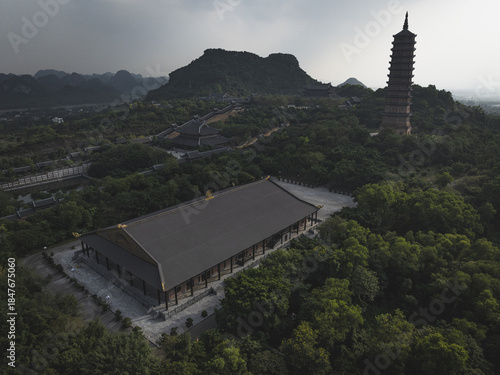 Aerial view of a pagoda nested amidst lush, green foliage, with a tall tower rising in the distance against a backdrop of rolling hills, Halong Bay, Vietnam.