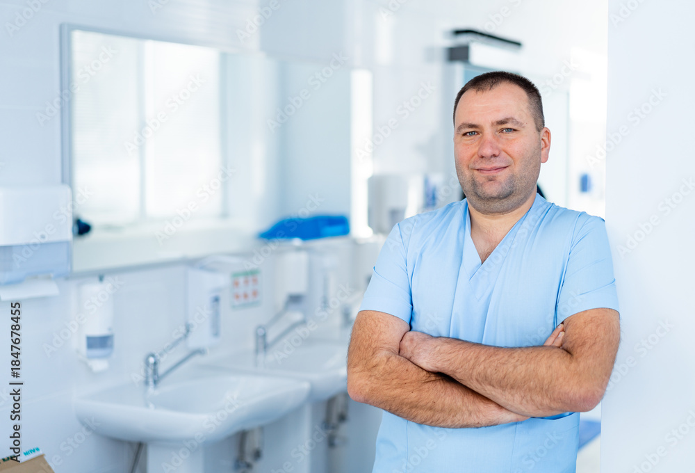 Obraz premium Confident Medical Specialist Standing with Crossed Arms. Waist-up portrait of a friendly male doctor in blue scrubs standing with arms crossed in a hospital examination room with a sink.