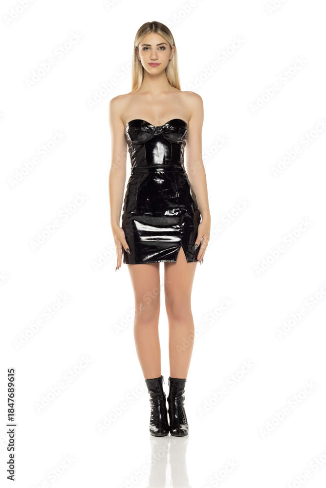 Fototapeta premium Full length studio shot of a blonde woman in a black latex mini dress and ankle boots standing confidently on a white background