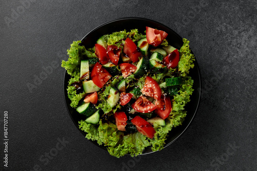 Vegetable salad of tomatoes, cucumbers, onions, lettuce dressed with aromatic dressing in a black plate on a gray background
