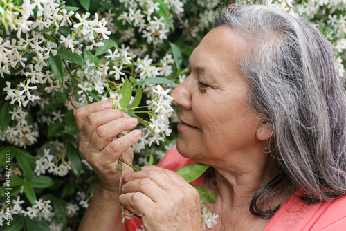 Close-up of a senior Latina woman smelling  jasmine flowers in a garden, enjoying a calm, peaceful moment outdoors.
