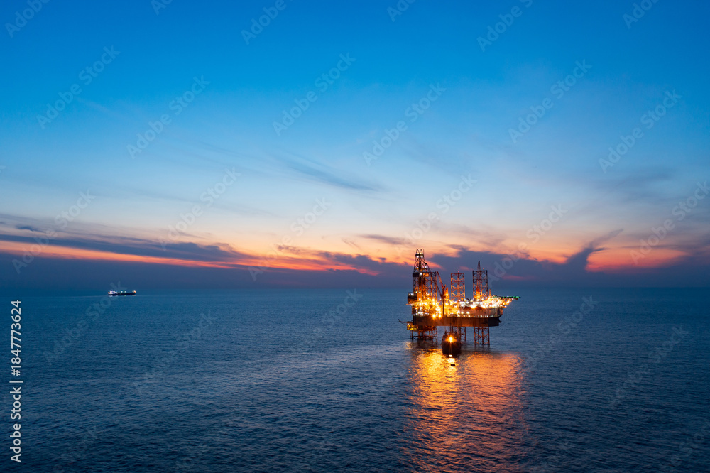 Fototapeta premium Aerial view from a drone of big jack up offshore drilling oil rig, production platform and supply vessel In the ocean during sunset - Oil and Gas Industry