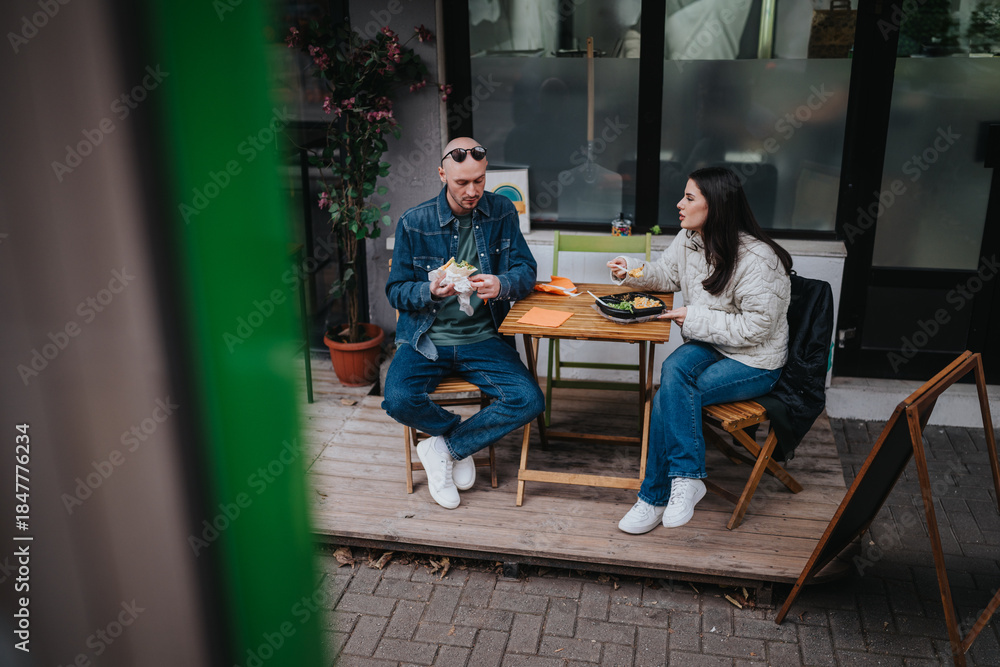 Fototapeta premium A man and a woman sit at a small outdoor cafe table, eating sandwiches. The casual scene shows friends enjoying a relaxed meal on a wooden patio.