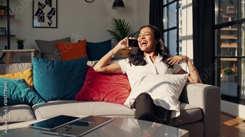 Young woman laughing during a relaxed smartphone call on a cozy living room sofa, enjoying sunlight, casual comfort and a connected, joyful moment at home