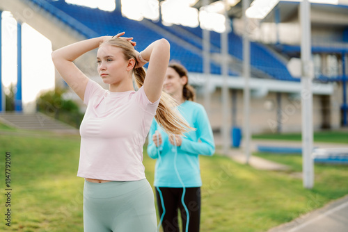 Two friends are enjoying a workout at the local track. One is stretching while the other prepares to jump rope. The warm sunlight casts a cheerful atmosphere on their exercise session.