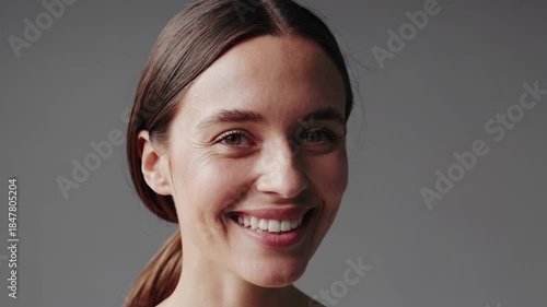 Young adult woman with bare shoulders smiling widely, showcasing natural beauty, healthy skin, and genuine happiness against a neutral grey background representing positivity and wellness