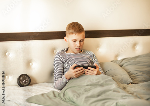 A young kid teenager boy sitting on a cozy bed, wearing striped pajamas, focused on his smartphone.