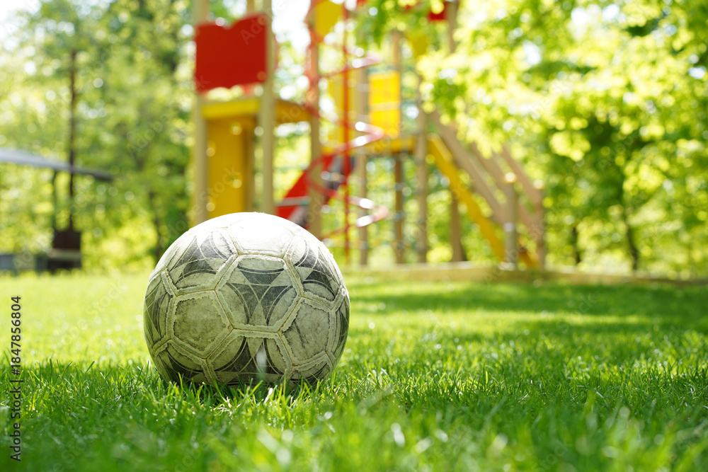 Fototapeta premium Old worn soccer ball sitting on vibrant green grass with a blurry colorful children's playground in the background.