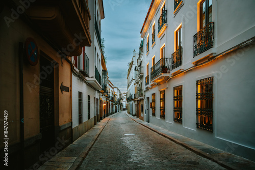 Empty narrow street with white buildings, wrought iron balconies, and glowing windows, capturing the serene evening ambiance of a historic Andalusian city in Cordoba, Spain