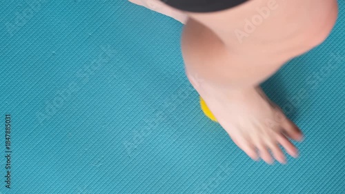 Female feet are doing foot exercises by rolling a prickly massage yellow ball under the foot on the yoga mat pressing down in different sides. Training to correct and relax the foot fascia. 4k footage