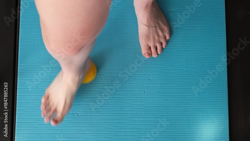 Female feet are doing foot exercises by rolling a prickly massage yellow ball under the foot on the yoga mat pressing down in different sides. Training to correct and relax the foot fascia. 4k footage