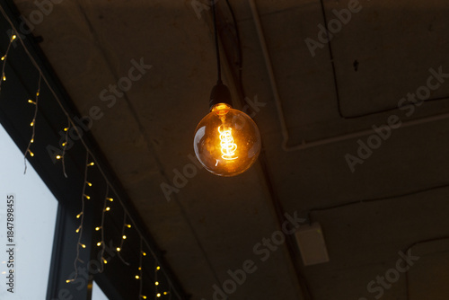 Single vintage bulb glowing near window, casting warm amber light on concrete ceiling and string fairy lights, creating intimate urban cafe atmosphere for latenight freelancer workspace, cozy