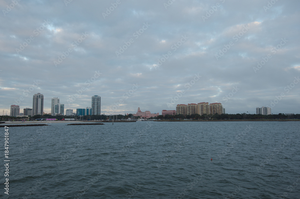 Obraz premium Coastal City Waterfront With Modern Buildings And Calm Vinoy Basin Under Cloudy Sky from the Pier in St. Pete, FL. Cityscape featuring modern high-rises along the shore, calm water, and a cloudy sky, 