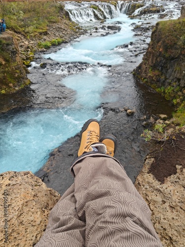 POV shot of a hiker sitting on a rocky cliff edge overlooking the vibrant blue Bruarfoss waterfall in Iceland. A first-person perspective of hiking boots and legs resting above the glacial river rapid