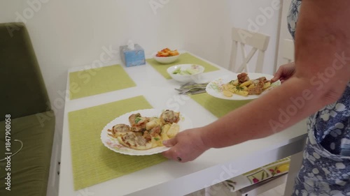 Woman bring fried meat with potato to table. Close up of hands holding white floral plate while serving fried meat rolls with potato slices from dark frying pan in modern home kitchen.