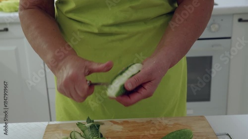 Close up of hands in yellow green apron peeling green cucumber with small knife, holding vegetable firmly while removing skin, preparing fresh ingredients for healthy meal at home.