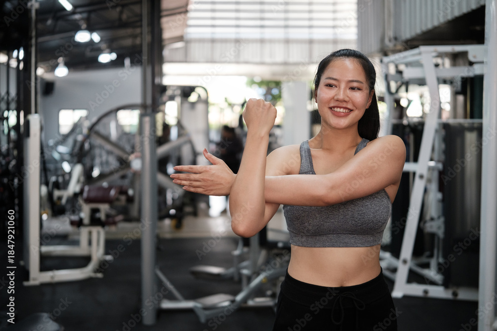 Obraz premium Smiling young woman stretching her shoulder in a modern gym before workout. Healthy lifestyle and wellness concept.