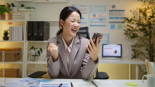 Excited businesswoman celebrating success while looking at her smartphone in the office. Concept of achievement and good news.