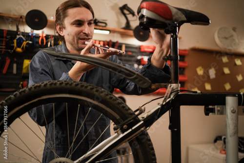 Fototapeta Professional mechanic recording voice message on smartphone while repairing bicy