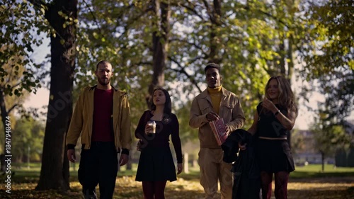 A diverse group of young friends takes a leisurely stroll through a park on a sunny autumn day. They chat, laugh, and share popcorn, enjoying each other's company amidst the vibrant fall foliage.