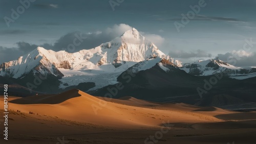 Fototapeta Naklejka Na Ścianę i Meble -  Snow-capped mountains rise behind golden sand dunes in a dramatic desert landscape at sunset