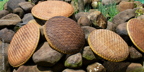 Five rustic, round Tampah or woven bamboo winnowing trays arranged on a textured, rough pile of dark volcanic rocks. Traditional Asian handicraft background.