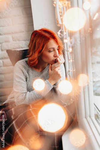 Woman with red hair sits by window during morning time and looks outside while lost in thought