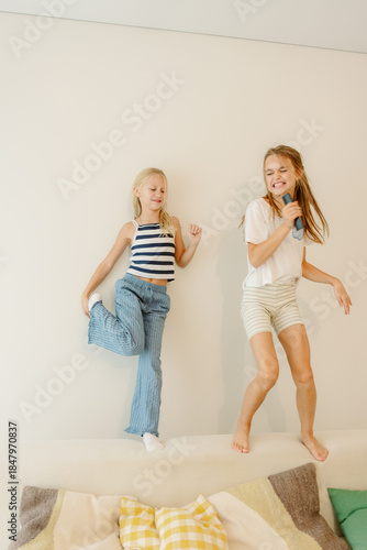 Two joyful children are having a great time in a cozy living room. One girl stands on the couch, dancing and singing into a microphone, while the other strikes a playful pose.