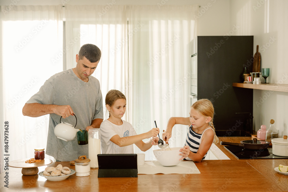 Fototapeta premium In a sunny kitchen, a father assists his daughters as they prepare ingredients for baking. Together, they mix and measure, sharing smiles and laughter in the cozy space.
