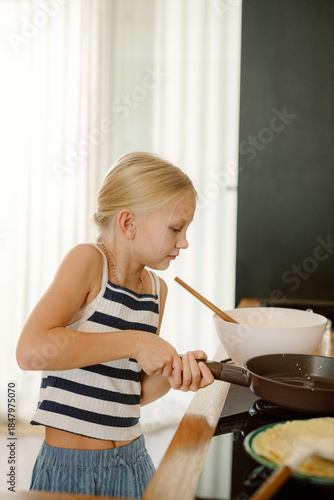 A young girl with a focused expression stirs a pan on the stove, preparing delicious pancakes. Natural light fills the warm kitchen, creating a cheerful cooking atmosphere.