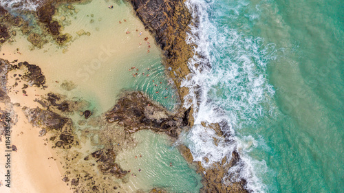 Aerial view of turquoise waves crashing against the rugged, dark rocks surrounding the sandy shores of Champagne Pools, Queensland, Australia.