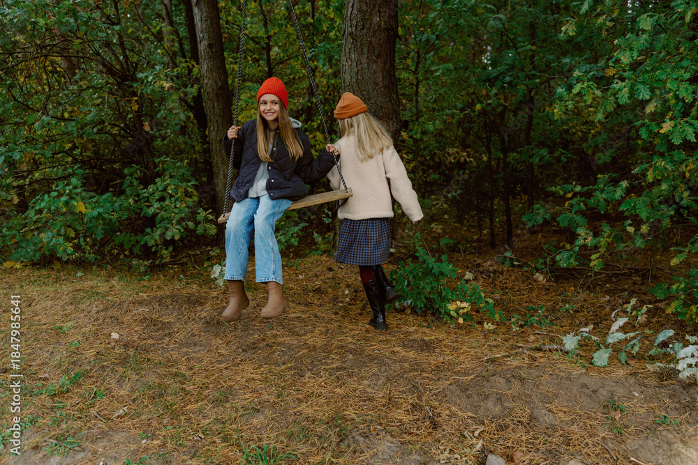 Fototapeta premium Two young girls swing joyfully in a peaceful forest. Surrounded by green trees and soft earth, they share laughter and friendship on a chilly autumn day.