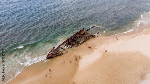 Aerial view of the rusting skeleton of the SS Maheno shipwreck stranded on the sandy beach with the ocean waves lapping, Fraser Island, Queensland, Australia.