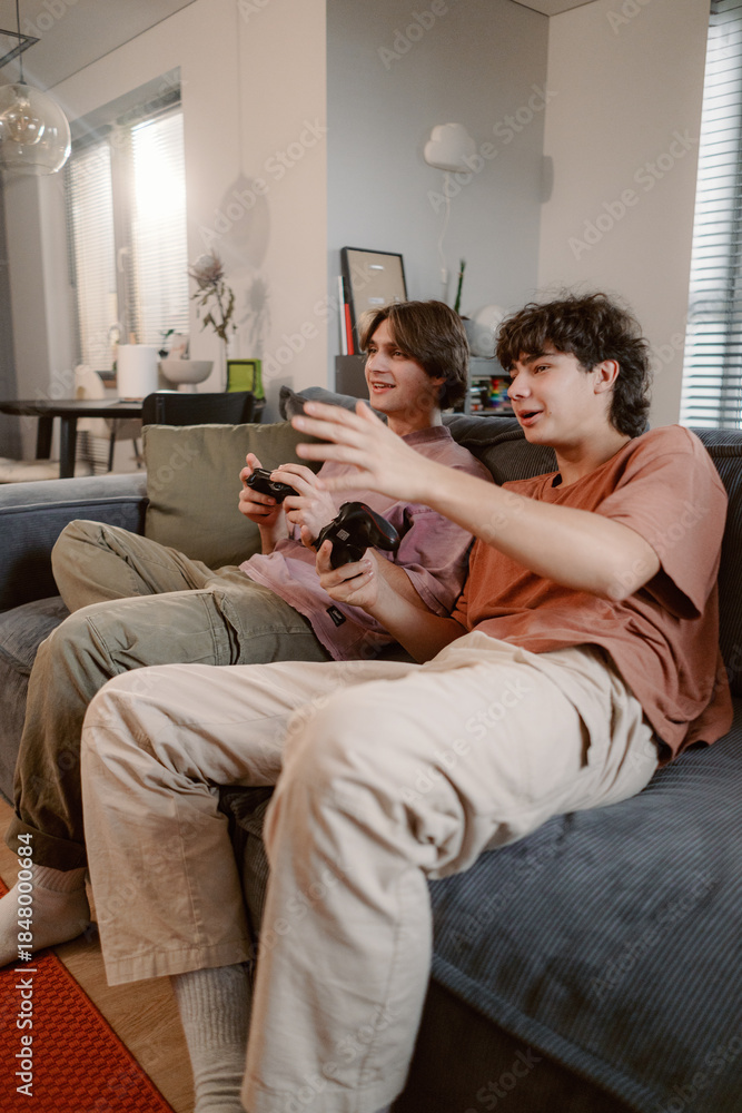 Naklejka premium Two friends sit comfortably on a couch, immersed in a gaming session. They share laughs and excitement as they play video games in a warm and inviting living room.