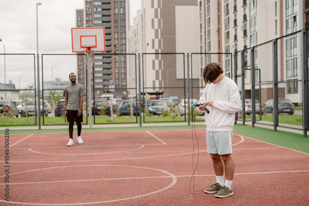 Fototapeta premium Two young men are having a relaxed time on a modern basketball court. One is checking his phone while the other waits, surrounded by urban buildings and parked cars.