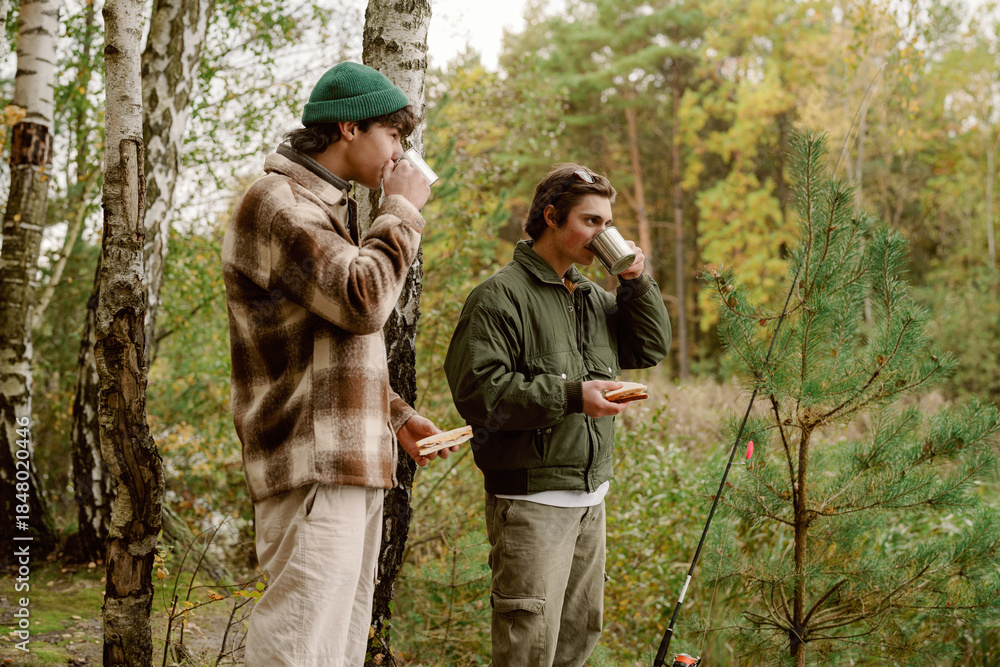 Naklejka premium In a peaceful forest, two friends sip hot beverages while savoring snacks, surrounded by tall trees and vibrant autumn colors. Laughter fills the air as they enjoy their time outdoors.